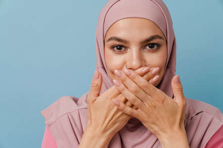 Young muslim woman looking at camera while covering her mouth isolated over blue backgroundの写真素材