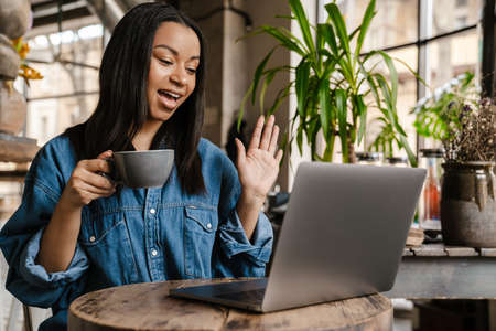 Smiling young african woman using laptop computer while sitting at the cafe table with cup of...