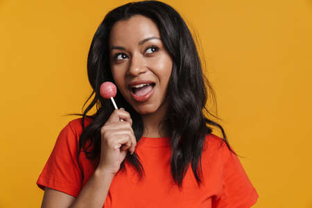 Young black woman smiling while licking lollipop isolated over yellow background