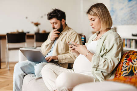 Bearded man and pregnant woman using cellphone and laptop while sitting on couch at homeの写真素材