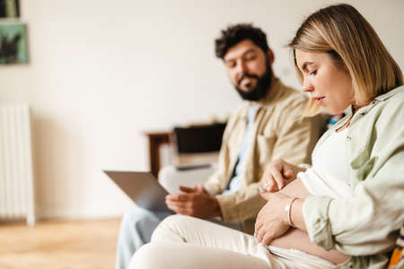 White woman touching her pregnant belly while sitting with her husband on couch at homeの写真素材