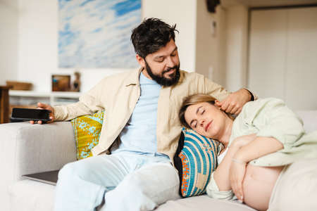 Bearded man caressing his pregnant wife while she sleeping on couch at homeの写真素材