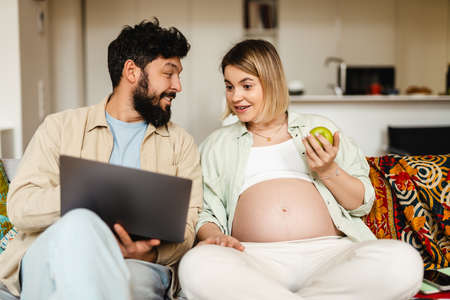 Bearded man and pregnant woman using laptop and eating apple while sitting on couch at homeの写真素材