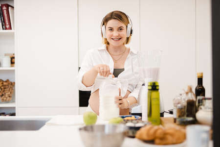 White pregnant woman in headphones making smoothy at homeの写真素材