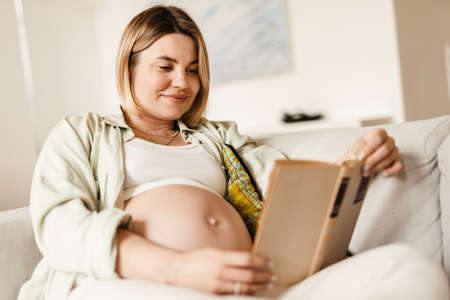 Young pregnant woman smiling and reading book while sitting on couch at homeの写真素材