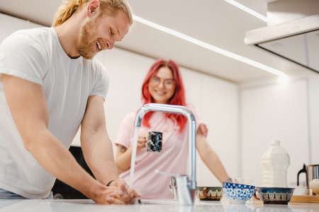 White man washing dishes and talking with his her girlfriend in kitchen at homeの写真素材