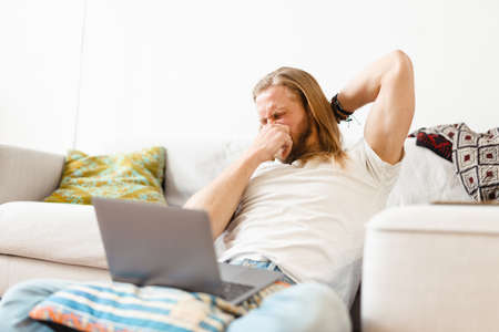 Bearded blonde man sneezing and using laptop while sitting on floor at home