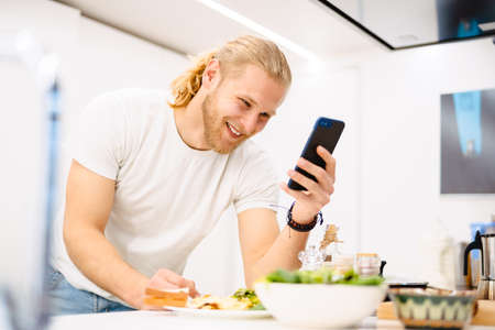 Young white man using cellphone while having lunch in kitchen at homeの写真素材