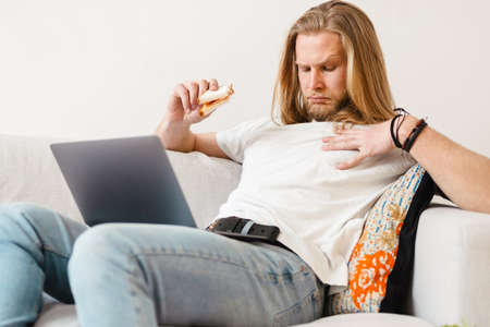 Bearded blonde man eating sandwich while working with laptop at home