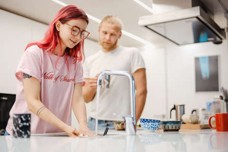 White man washing dishes and talking with his her girlfriend in kitchen at homeの写真素材
