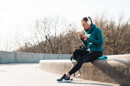 Young smiling sportswoman in headphones with prosthesis sitting on a fitness mat using mobile phone outdoorsの写真素材