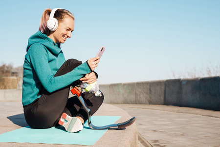 Young sportswoman in headphones with prosthesis sitting on a fitness mat using mobile phone outdoors