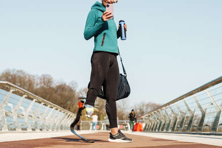 Cropped image of a young healthy sportswoman with prosthetic leg standing holding mobile phone and...