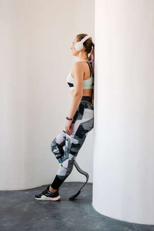 Young sportswoman with prosthesis in headphones leaning on wall indoors