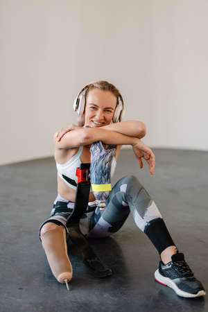 Disabled happy young white sportswoman in headphones with prosthetic leg sitting on a floor in room
