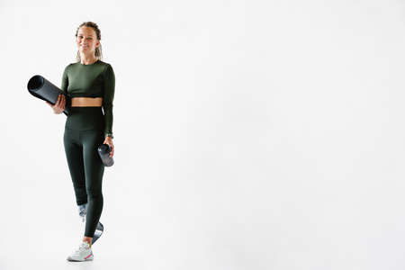 Young sportswoman with prosthesis holding fitness mat and water bottle isolated over white wall