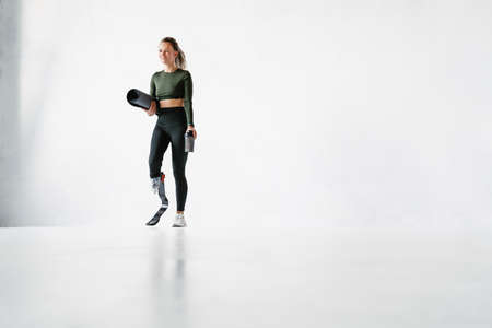 Young sportswoman with prosthesis holding fitness mat and water bottle isolated over white wall