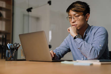 Focused asian guy in eyeglasses working with laptop while sitting at table in office