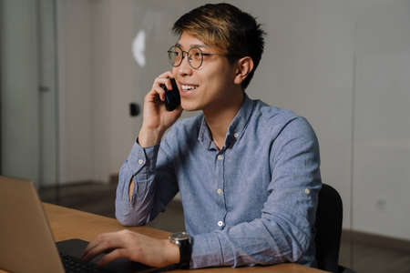 Happy asian guy talking on mobile phone while working with laptop in office