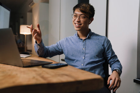 Smiling asian guy in earphones gesturing while making video call on laptop in officeの写真素材