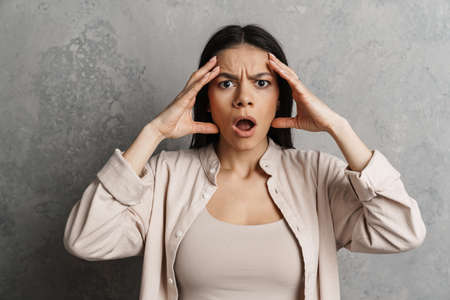 Brunette hispanic shocked woman holding her head and looking at camera isolated over grey wall