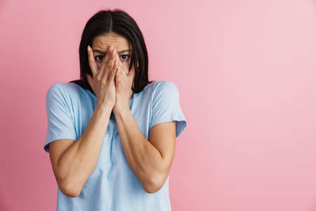 Young hispanic scared woman covering her face and looking at camera isolated over pink wall