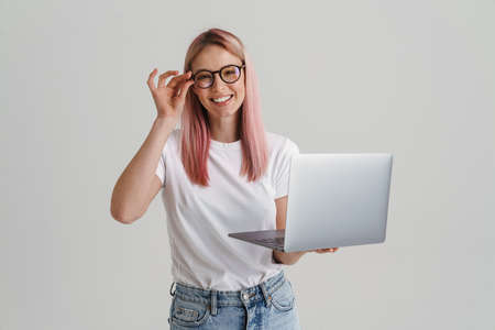 Happy young blonde white woman in glasses standing holding laptop computer over gray wall backgroundの写真素材