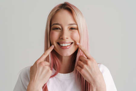 Young woman with pink hair smiling and pointing fingers at her cheeks isolated over white backgroundの写真素材