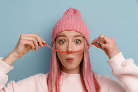 Young white woman with pink hair having fun while holding belt candy isolated over blue wall