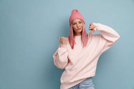 Young woman wearing pink hat frowning and showing thumb down isolated over blue wallの写真素材