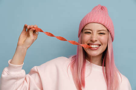 Young white woman with pink hair smiling and eating strip candy isolated over blue wallの写真素材