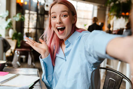 Young beautiful woman with pink hair taking selfie photo while studying in cafe
