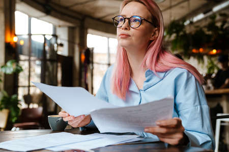 Young beautiful woman with pink hair doing paperwork while sitting in cafeの写真素材
