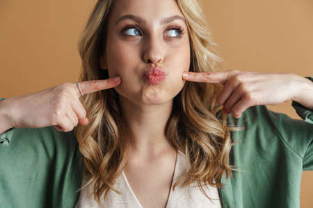 Amusing beautiful woman grimacing and pointing fingers at her cheeks isolated over beige wallの写真素材