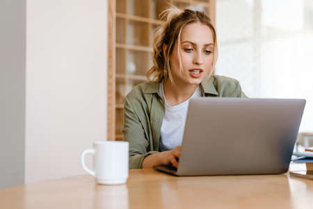 Young white woman using laptop while sitting at table in room