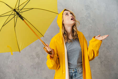 Lovely excited young woman wearing waterproof coat holding umbrella isolated over gray background