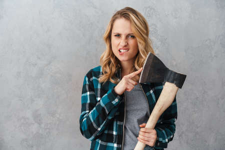 Angry beautiful woman holding and pointing finger at hatchet isolated over gray wallの写真素材