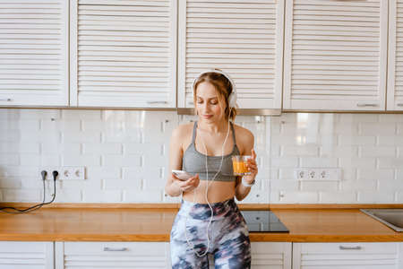 Smiling young white woman in sportswear drinking juice listening to music with headphone in the...