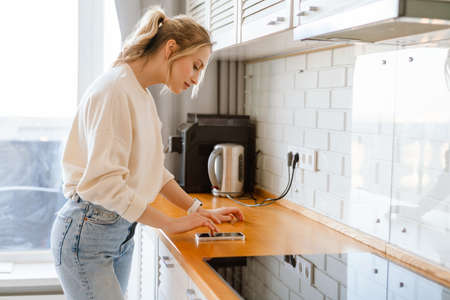 Young woman using mobile phone while standing in kitchen at homeの写真素材