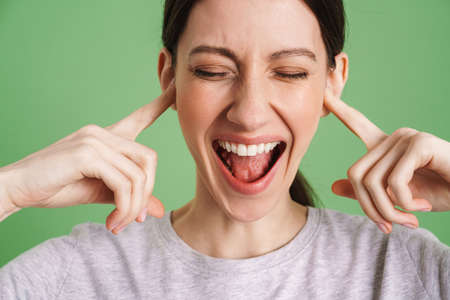 Young brunette woman screaming while plugging her ears isolated over green background