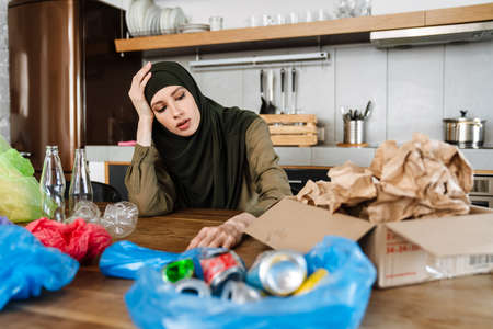 Young muslim woman wearing hijab separating waste while sitting at table in kitchen