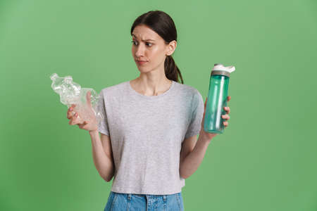 Young brunette woman showing disposable and reusable bottles isolated over green backgroundの写真素材
