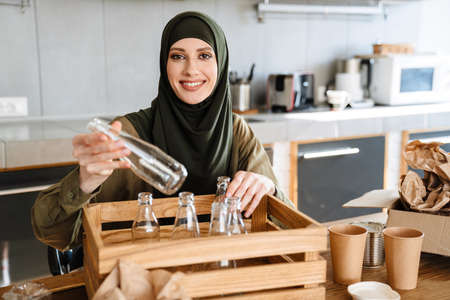 Young muslim woman wearing hijab separating waste while sitting at table in kitchenの写真素材