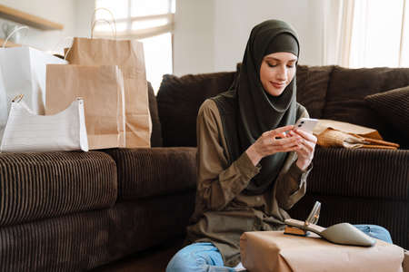Young muslim woman using cellphone while sitting on floor with shopping bags at homeの写真素材