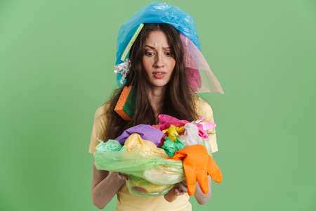 Young brunette woman with plastic waste expressing surprise at camera isolated over green background