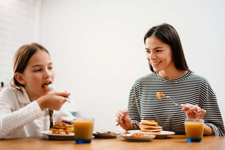 Cheerful beautiful mother and daughter smiling while having breakfast in cozy kitchenの写真素材