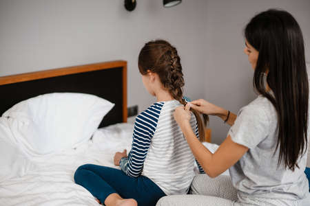 Nice beautiful mother making hairstyle to her daughter while sitting on bed at home
