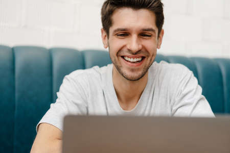 Smiling young man student sitting in cafe indoors with laptop computer, looking at screenの写真素材