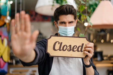 Young man waiter in apron and face mask showing closed sign while standing in cafe, outstretched handの写真素材