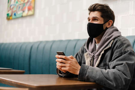 Young brunette man wearing medical mask sitting at the cafe table indoors, holding mobile phone